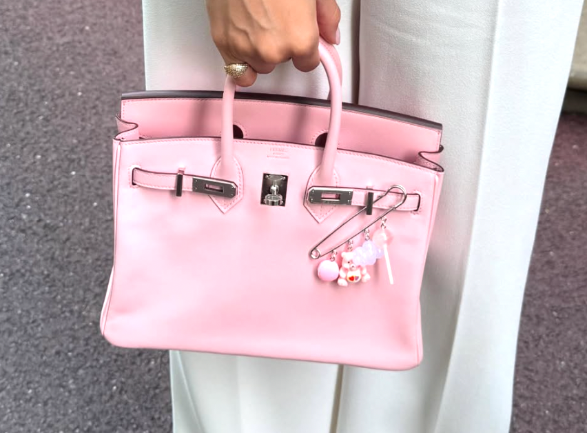 Pink handbag with a decorative charm held by a person wearing white pants.